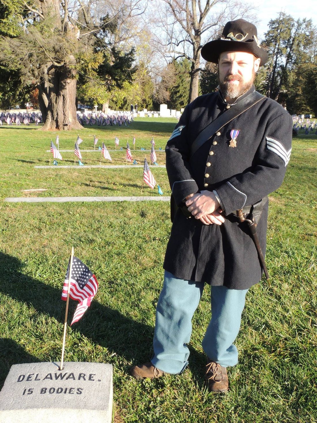 Sean Protas in Union uniform at Gettysburg National Cemetery, beside a stone marker reading Delaware, 15 bodies